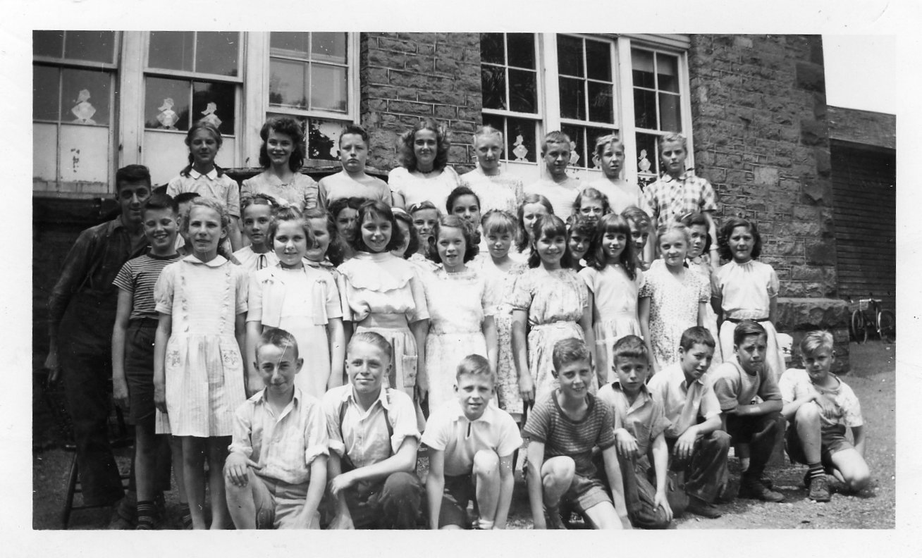 Black and white photograph of a group of children posing on the south side of Victoria School. Two banks of windows behind and frame addition to the east of the building with a bicycle leaning against it. There are four rows of students. The first row is kneeling, the second two rows are standing, the third row is standing (presumably) on a bench. 