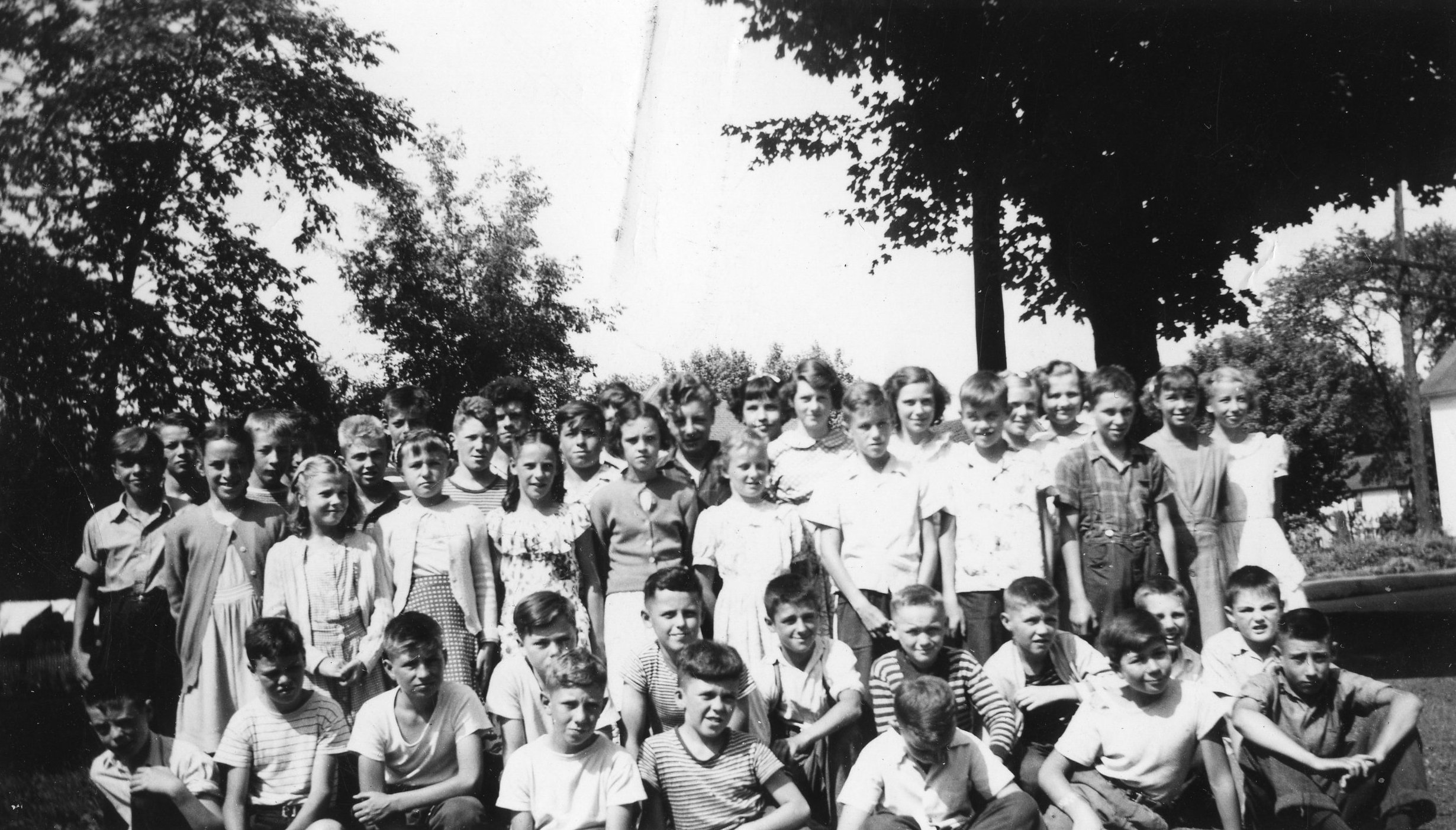 Greyscale photograph featuring children lined up and arranged into three rows for a class photo. First row sitting on grass, second row kneeling, all behind standing.