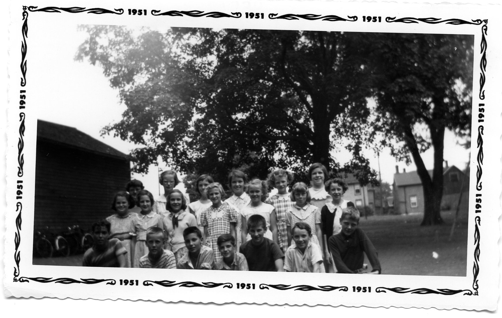 Black and white photograph of a class portrait outside Victoria School. From Robert C.M. Allen. There is a border around the edge of the photograph with the year 1951 inscribed in it. There are three rows of students, both boys and girls. To the left is a wooden shed with two bicycles against it.