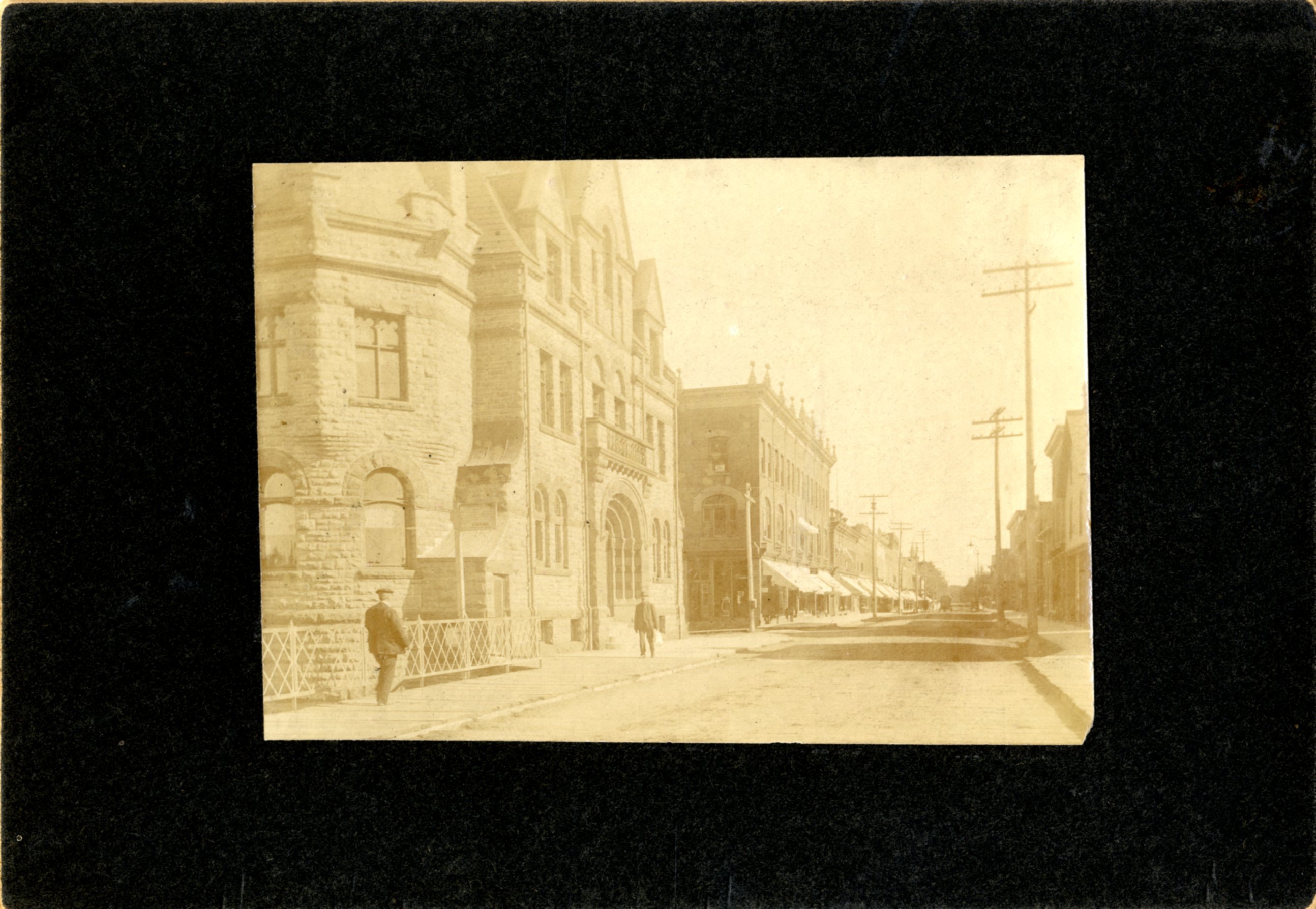 Sepia photograph mounted on black mat. Photo of Bridge Street, Carleton Place, taken from bridge looking south. Bridge railing, Town Hall, Taylor's, dirt road, wooden sidewalks, awnings, telephone poles, two pedistrians, two buggies in street.