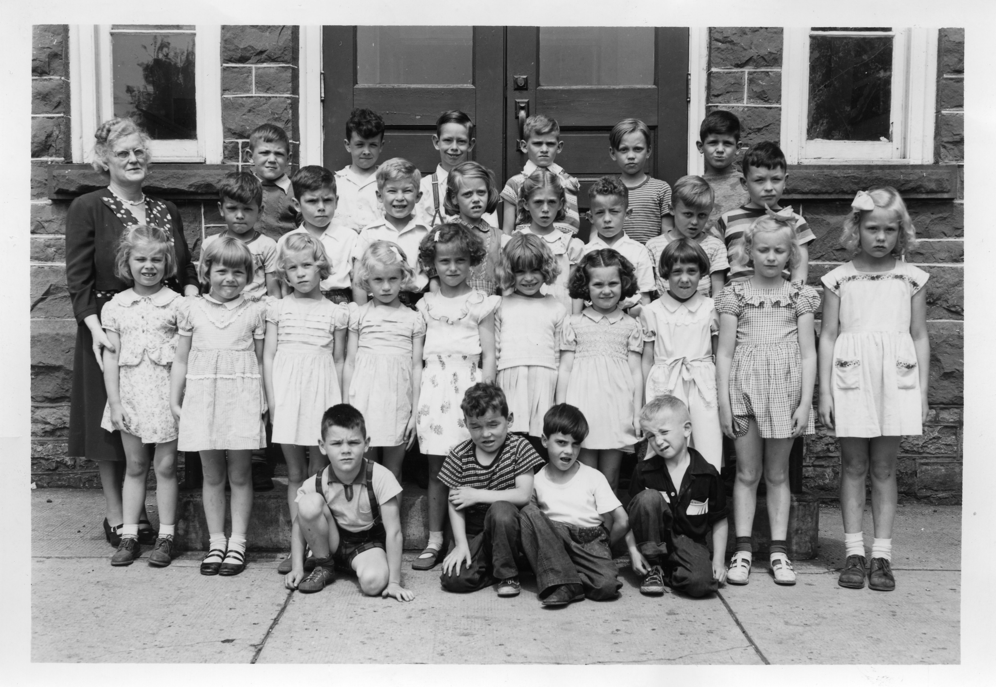 Black and white photograph of a class portrait taken outside Victoria School. The photograph is taken on the front steps of the school, with the front doors and steps visible. There are four rows of children. The front and back rows are boys, the two middle rows are girls. The teacher is Miss Olive Robertson,standing to children's right. Stamped in black on reverse: "Ernie Foote, Photographer, Box 589, Carleton Place". 