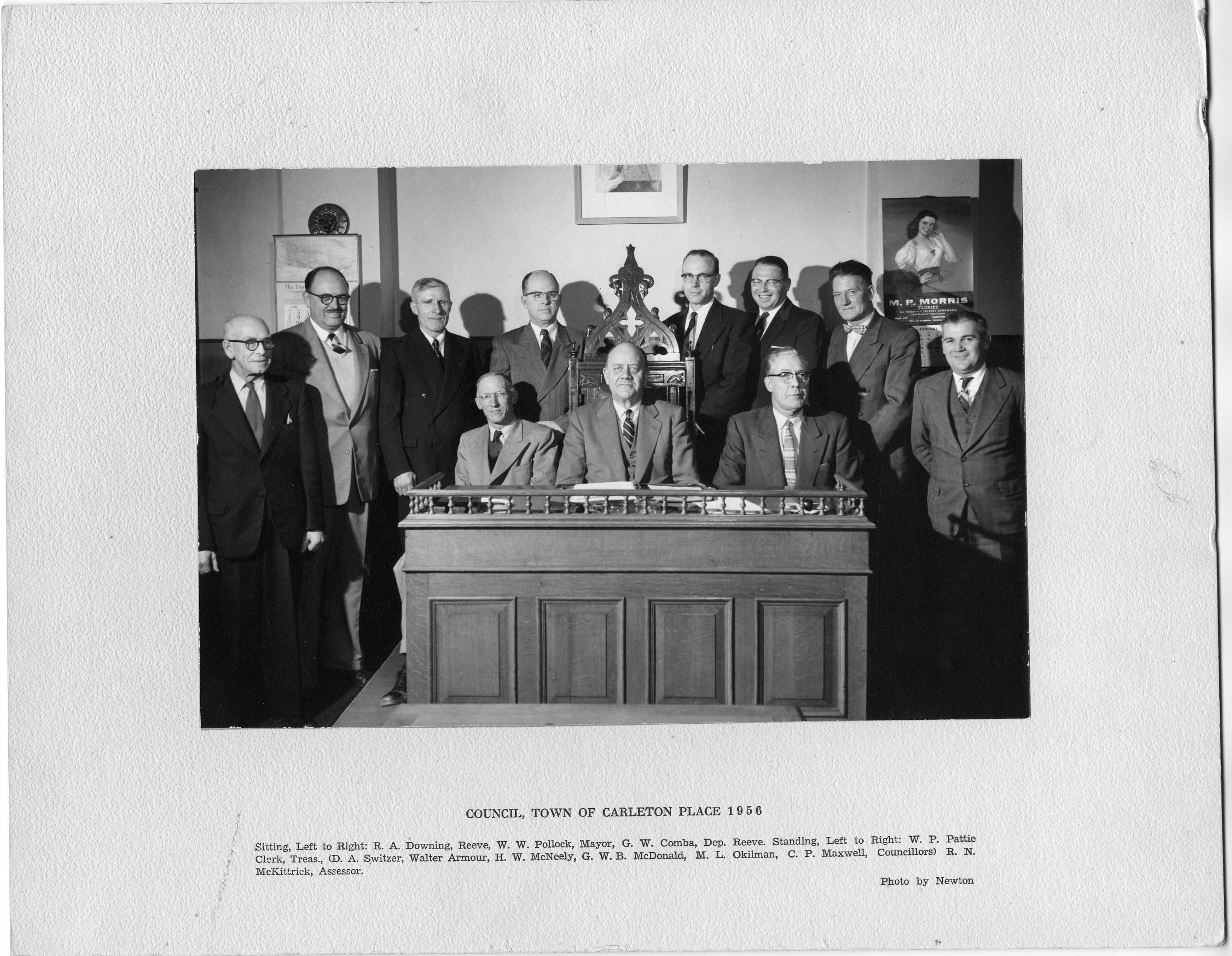 A black and white photograph mounted on white mat. "Council, Town of Carleton Place 1956". There is one group of men behind and there are three men sitting at a desk. The mayor is seated in an ornate chair. Two calendars are at either side of the room. Calendar on the right is for "M.P. Morris, Florist." Printed on mat: "Sitting, Left to Right: R.A. Downing, Reeve, W.W. Pollock, Mayor, G.W. Comba, Dep. Reeve. Standing, Left to Right: W.P. Pattie Clerk, Treas., (D.A. Switzer, Walter Armour, H.W. McNeely, G.W