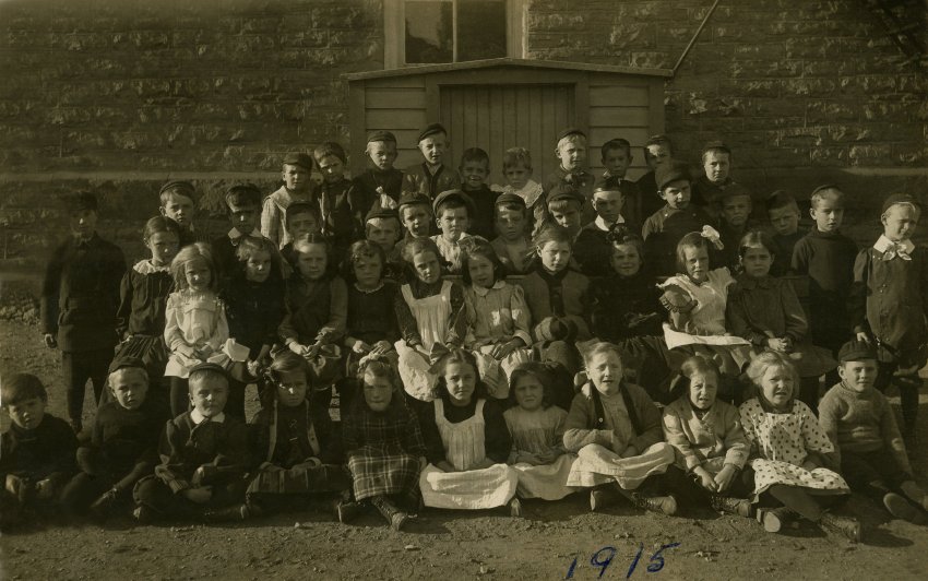 Black and white photograph of school children behind Victoria School. The class portrait has four rows, both boys and girls. The first row is seated. Behind them is a wooden shed. At the bottom is "1915" written in blue ink.