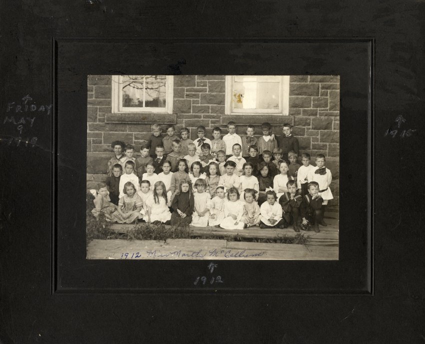 Black and white photograph on a black mat of young school children lined up in rows in front of the brick school. 