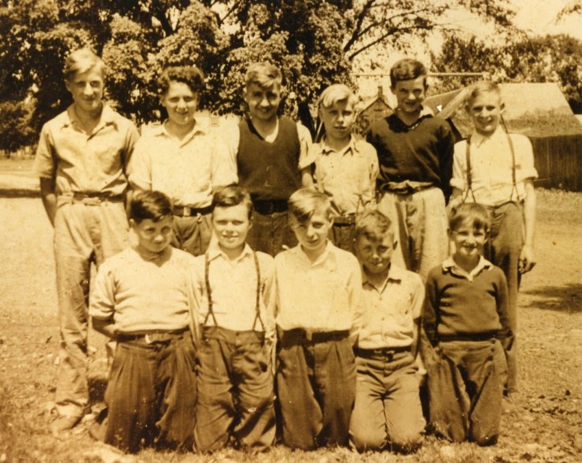 Sepia-toned reprint of black and white photograph of the Grade 7 boys at Victoria School. There are two rows, the top is standing and the bottom is kneeling. The boys have their hands behind their backs. The photograph was taken in the yard of the school. A wooden shed and roof of a house are visible in the back.