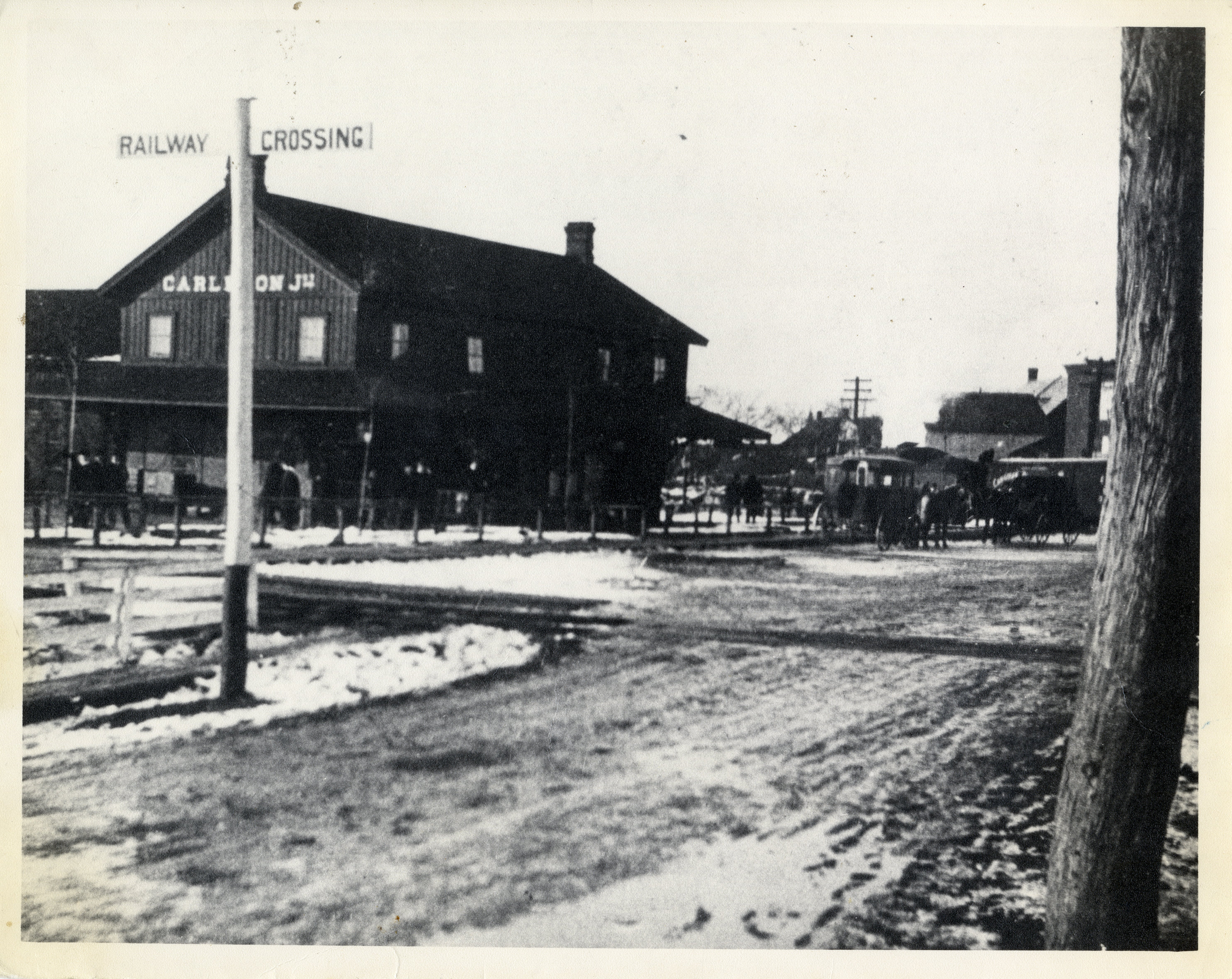 Black and white photograph of the old Carleton Place train station. It is visible in the upper left hand quadrant of the photo, behind a T-shaped sign reading "railway crossing". The right hand side of the photo is taken up by an empty street.