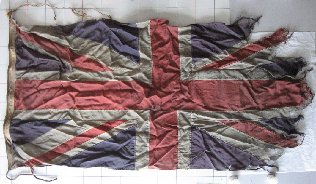 Oversized Royal Union (or Union Jack) flag. Colours are quite faded (blue, white, and red). It is mounted onto a lightly padded rigid support backing and reinforced with a wood frame. At its right edge are newly added dyed fabrics to support, match, and complete the flag. 