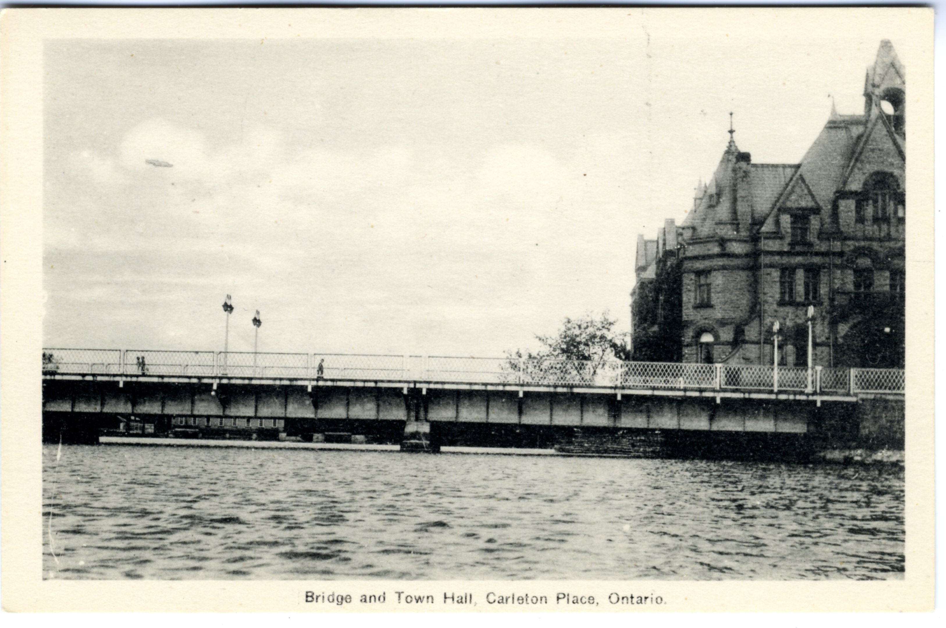 Rectangular postcard with black and white photograph of "Bridge and Town Hall, Carleton Place, Ontario". Shows bridge over Mississippi River and portion of Town Hall to right. Three people walking over bridge. Stone building visible in background behind bridge to left. Unused. Printed on reverse: "The Photoflatine Engraving Co. Ltd., Toronto".