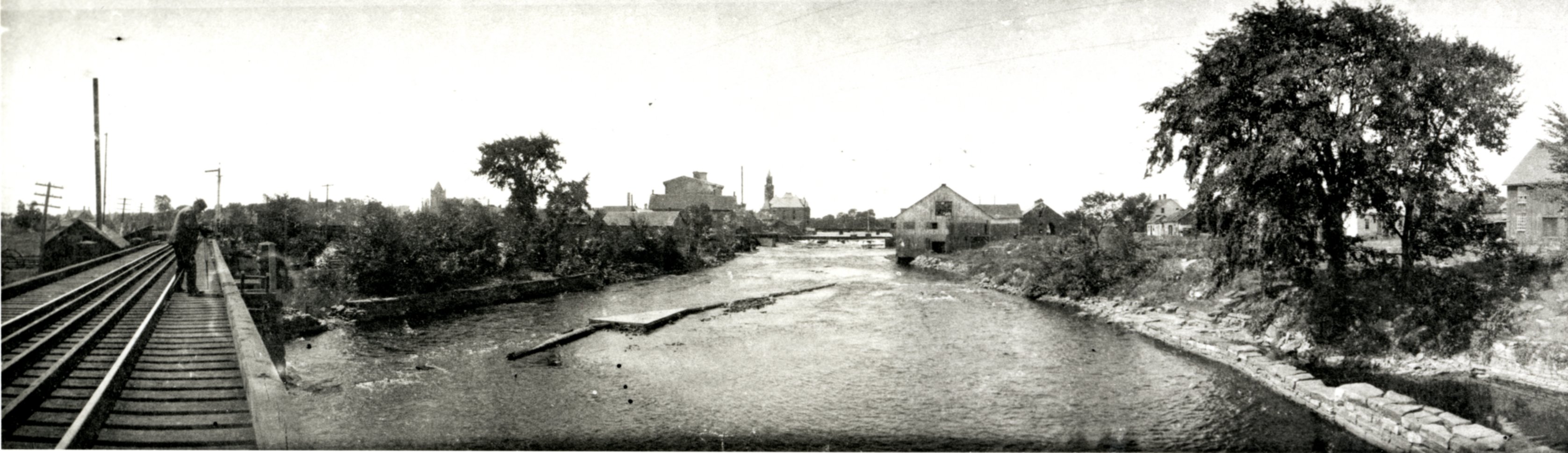 Black and white reprint of large panorama photograph taken from railway bridge across Mississippi River in Carleton Place circa 1905. Photograph features photographer standing on bridge, the Methodist Church, Boulton-Brown Grist Mill, the Town Hall, Gillies and McLaren Saw Mill (Canada Lumber Company), and a large elm tree. Photograph also shows how water in river was diverted for the mills. Original is found at National Archives C-001343.