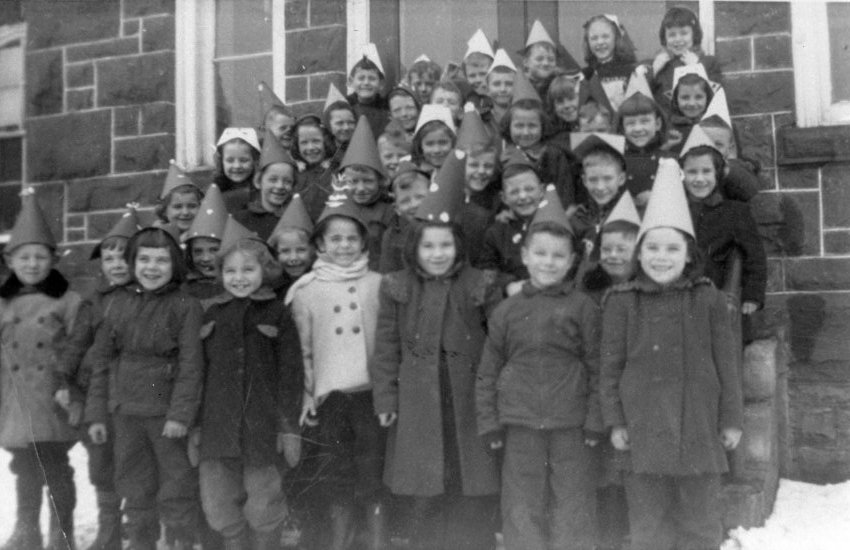 Black and white reprint of a grade 1 class on the front steps of Victoria School. The photograph is angled at the right of steps. It is winter. There are many children on the steps wearing paper cone hats with decorations. Presumably "elf" hats.