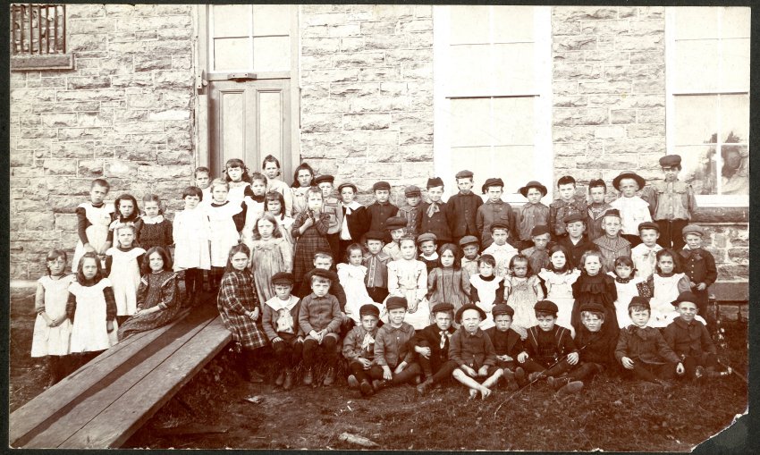 Black and white photograph on black mat. Photograph shows a large group of children (~60), approximately 6 or 7 years old, standing behind Victoria School. Girls are wearing dresses, mainly plaid, many with white aprons over the front. Boys are generally wearing dark button-up jackets with page-boy caps. There is a long wooden ramp up to the back door of the school. In the top-left corner of the photograph, the bricked-in barred window from the jail (lock-up) can be seen.