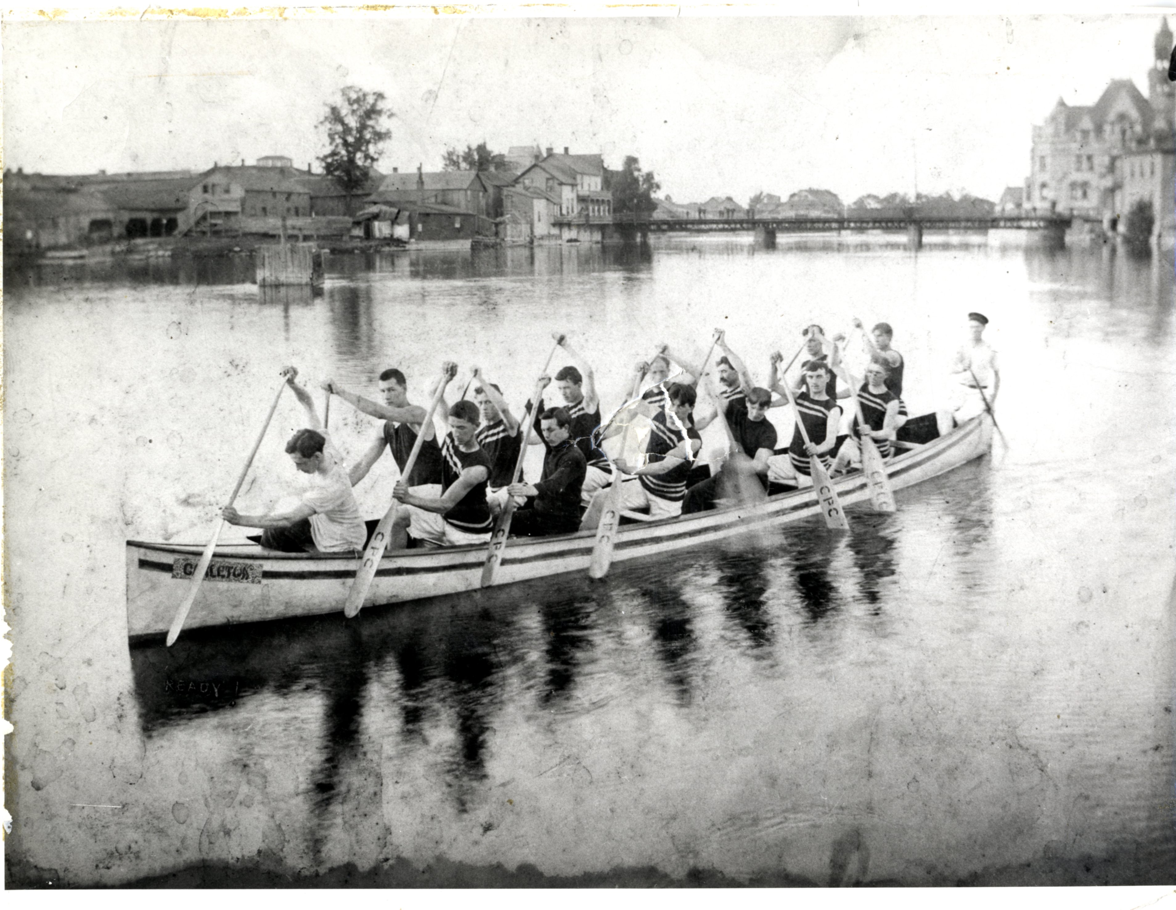 A picture of 15 men in a war canoe, 1905. The men are wearing sleevelss uniforms with two stripes across the chest and waist. The man at the back is wearing a hat. Their paddles are at the ready. In the background are the bridge and Town Hall. On the left side of the river are Findlay buildings. There are also buildings that are on Bridge Street. National Archives number C 29322. There is a larger copy mounted on cardstock available. It is stored in the box Oversize - Canoe Club Photographs