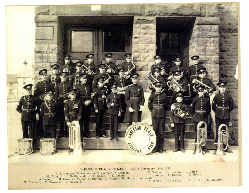 Black and white photograph of The Carleton Place Citizens Band. They are lined up on the side steps of the town hall. Each member is holding his instrument and dressed in uniform. They are lined up in four rows. Bass drum is front row centre. Signature of E.V Godwin is in the bottom right corner. Reprint. 