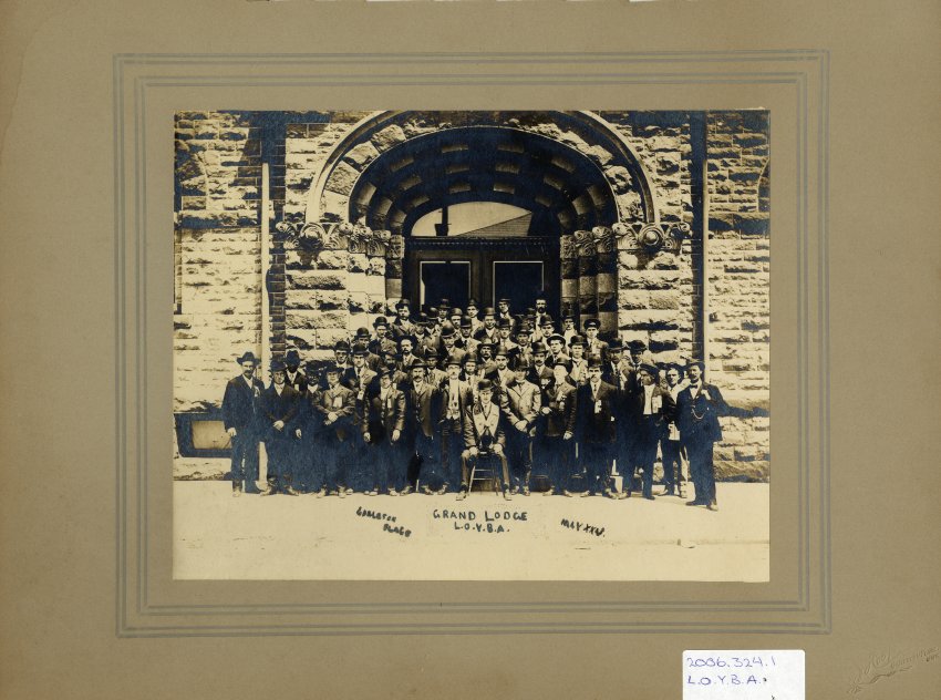 Black and white photograph mounted on beige mat. A portrait of the Grand Lodge. The men are situated on the front steps of the Carleton Place Town Hall. . The men are all wearing suits and are all wearing hats. One man who is front and center is seated upon a stool. Some of the men also have medals over there breast pocket. Caption at the bottom of the photograph reads "Carleton Place Grand Lodge L.O.Y.B.A. May XXV".