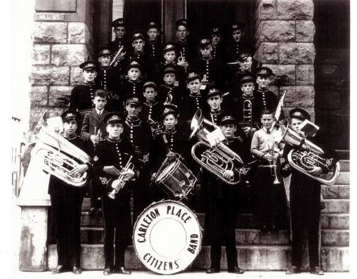 Black and white reprint of a photograph of the Carleton Place Citizens' Band. The band consists of 26 boys and men, all in uniform. The band is standing on the front steps of the Town Hall in Carleton Place. A sheet of names, provided by Jack Peckett accompanies photograph (see history). * Foamcore copy available.