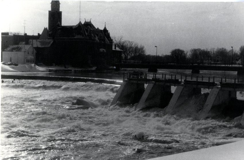 A black and white photograph of the dam and the town hall. The water is pouring over the dam. There is snow on the far bank of the river. The bridge can be seen. The photograph has a very dark contrast.