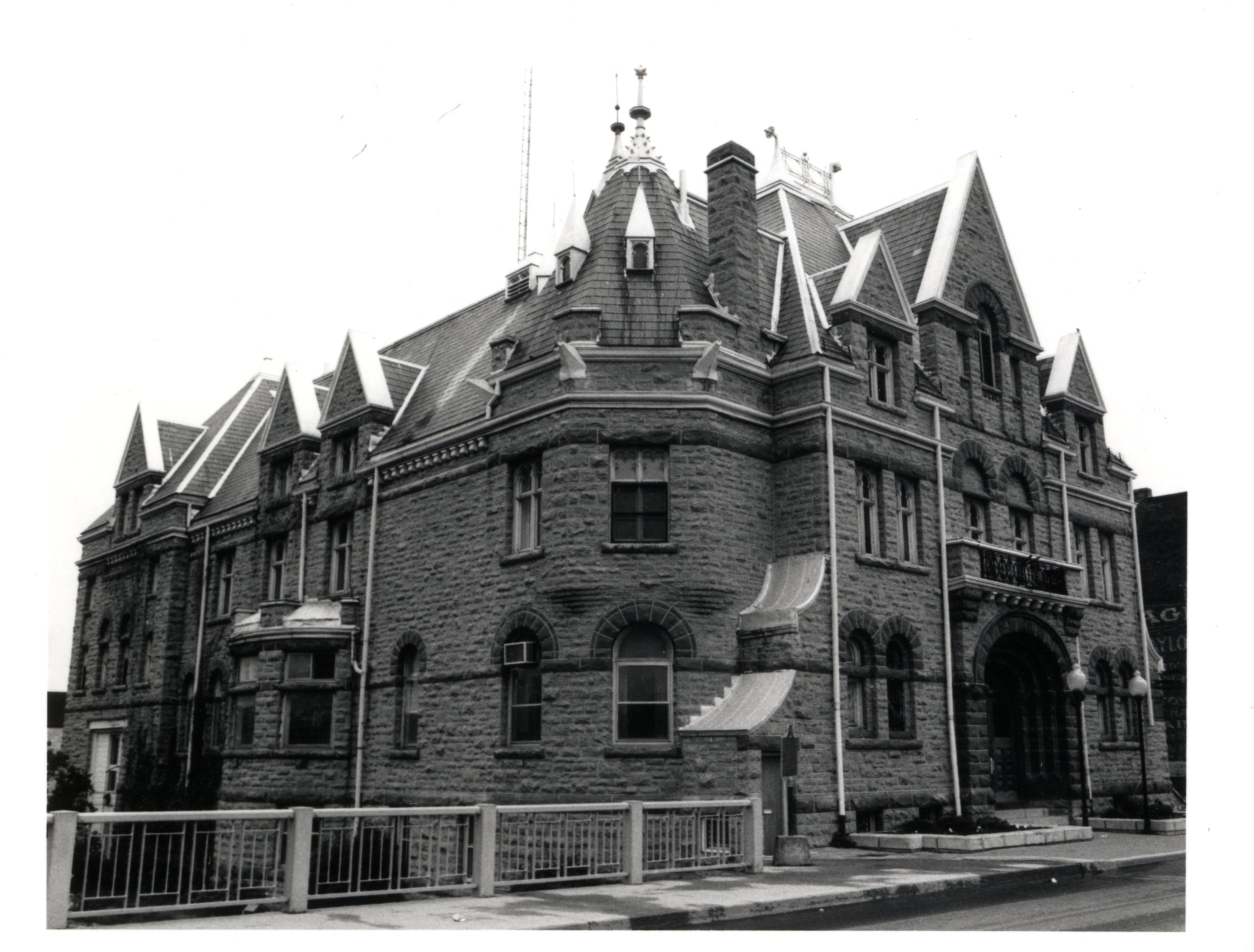 A black and white photograph of the town hall in Carleton Place. The picture is taken of the North-West corner of the town hall. Part of the bridge railing can be seen. A sign for Taylor's Hardware can be seen in the farright of the photograph.