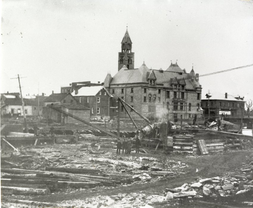 Black and white photographic reprint (c 5362) from National Archives of Canada showing view of Mississippi River during the construction of Horace Brown and Sons dam, Munro and McCuan, contractors. The Town Hall and other buidings are in the background. Pattersons Furniture Store is visible to the right of Town Hall. Hawthorne Mill is also visible to the very right. There are men standing in the river where there is no water. In the middle of the river is (what looks like) a cement mixer. There are logs all