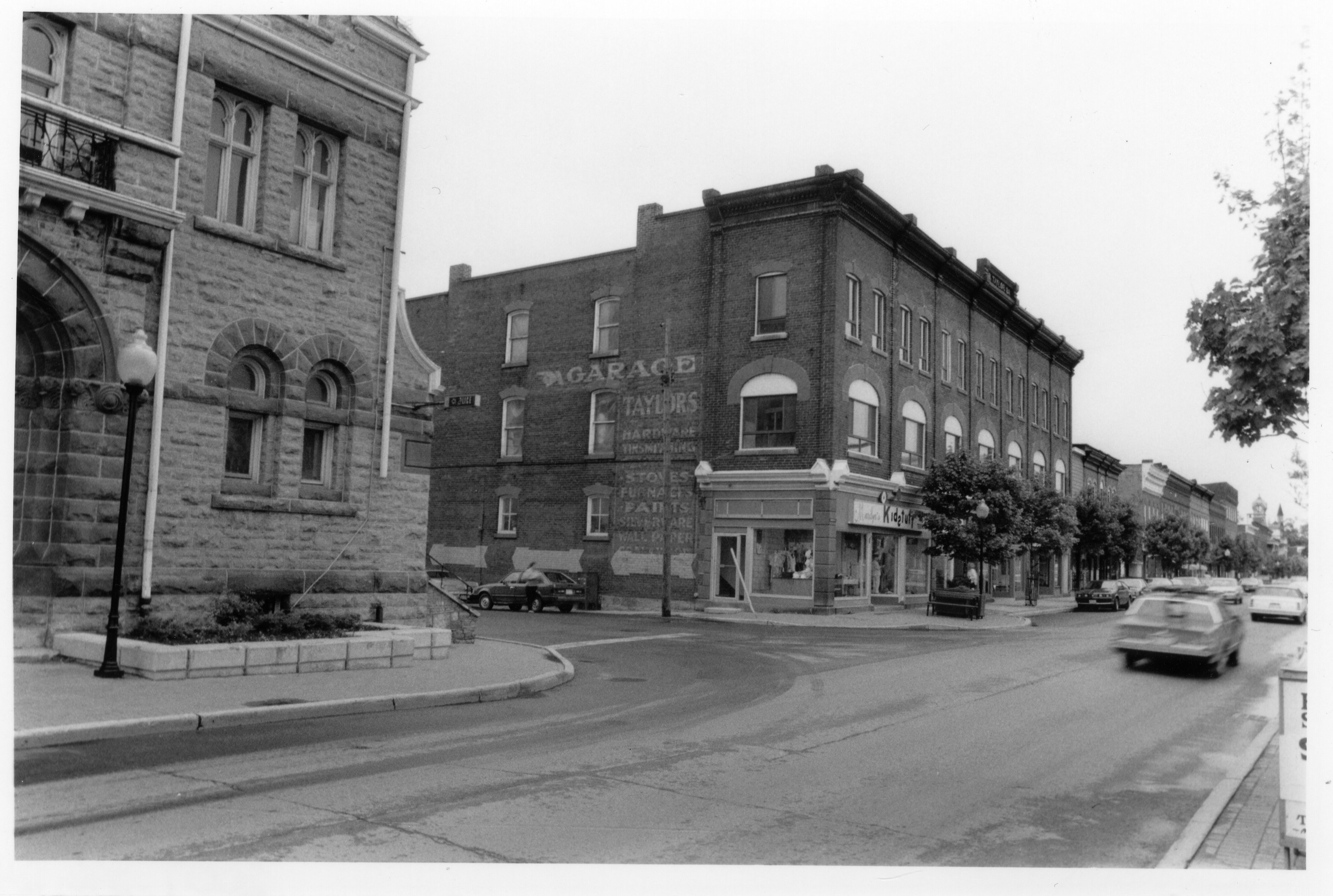 Photograph of the Kids Turf children's clothing store on the corner of Bridge and Mill street , taken from in front of the Town Hall. The large Garage, Taylors Hardware sign painted on the side of the wall is still visible. Part of a lot of black and white photographs of the East side of Bridge Street around the 1980's.