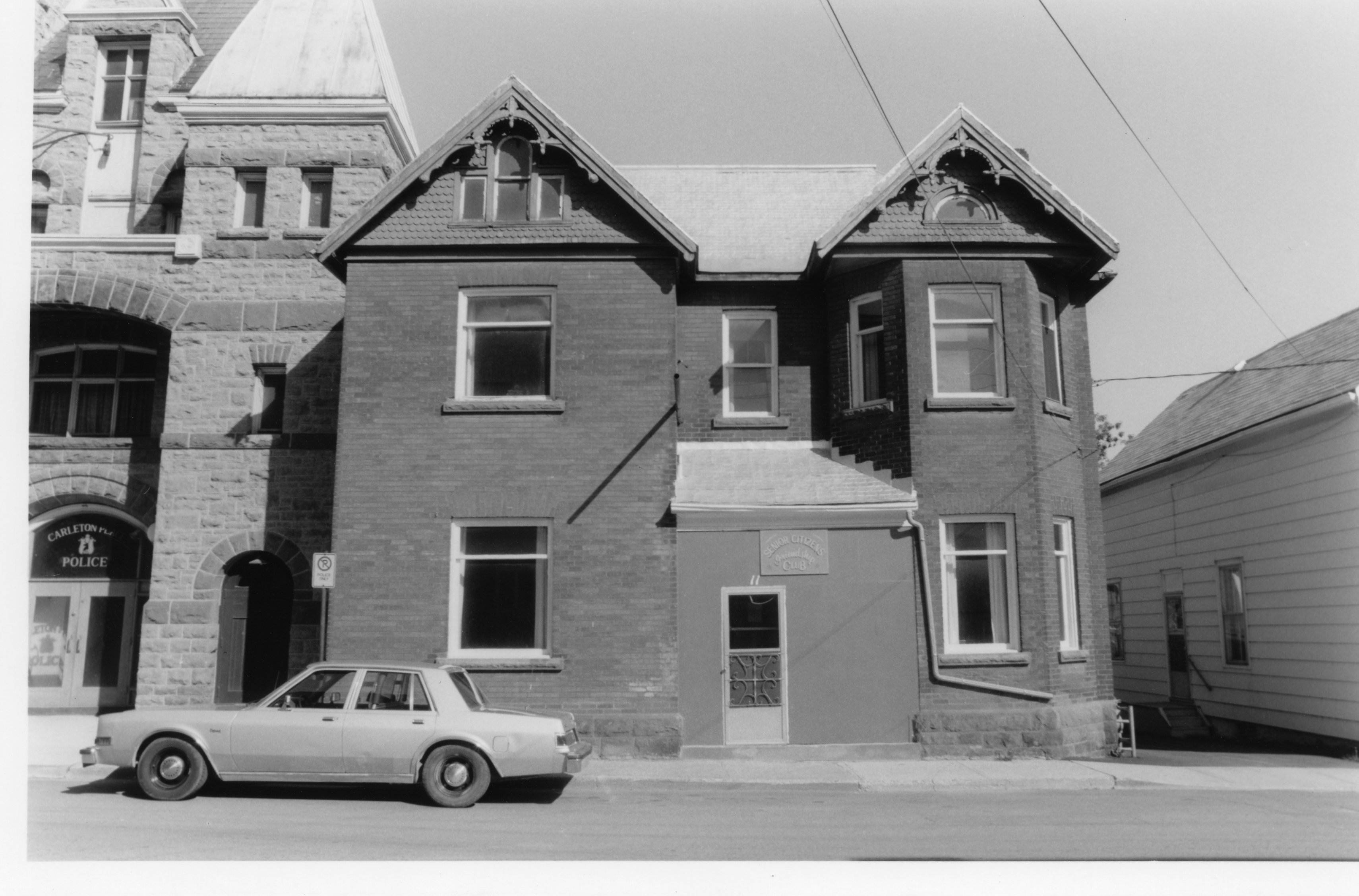 Black and white photograph of a large brick house on Mill Street next to the Town Hall. Above the front door is a sign that reads "Senior Citizen's Friendship Club". Parked in front is a silver car. To the left of the building the Town Hall is visible. In the window that can be seen is a sign for the Carleton Place Police Department. Part of a lot of black and white photographs taken of Mill Street in the 1980's.