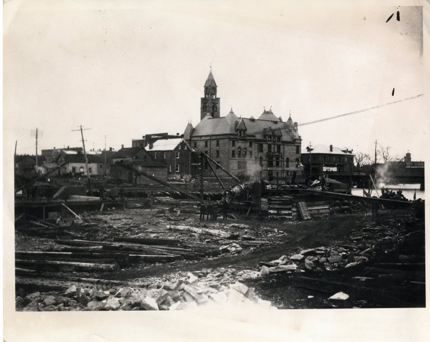 Black and white photographic print (c 5362) from National Archives of Canada showing view of Mississippi River during the construction of the H. Brown and Sons dam in 1907. Munro and McCuan, contractors. The Town Hall and other buidlings are in the background. Pattersons Furniture Store is visible to the right of Town Hall. Hawthorne Mill is also visible to the very right. There are men standing in the river where there is no water. In the middle of the river is (what looks like) a cement mixer. There are l