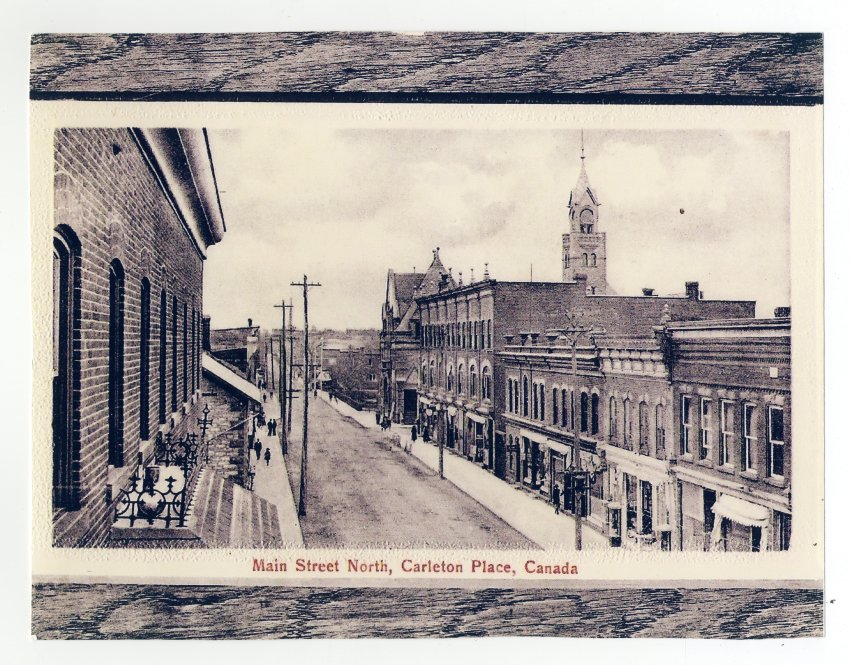 A black and white postcard from Carleton Place. The postcard is of the Main St. North. The photo is taken from the balcony of the Queen's hotel. The bridge can be seen in the background as well as the tower of the Town Hall. Taylor's block and be seen on the right. Written at the bottom "Main Street North, Carleton Place, Canada."