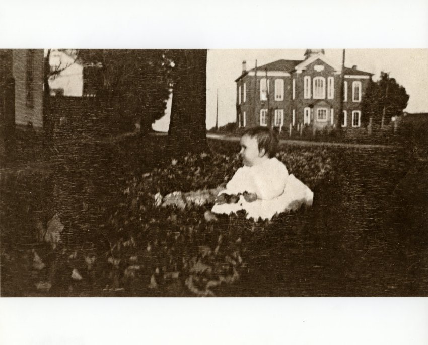 Sepia-toned photograph of a young child in a white dress, sitting on the grass. Victoria School is visible in the back right. A house is at the left edge. There is a tree behind the child.