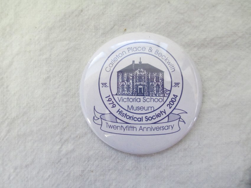 White pin with blue logo and lettering: "Carleton Place & Beckwith Historical Society 1979 - 2004, Twenty-fifth Anniversary, Victoria School Museum". There is an illustration of the museum building in the center..