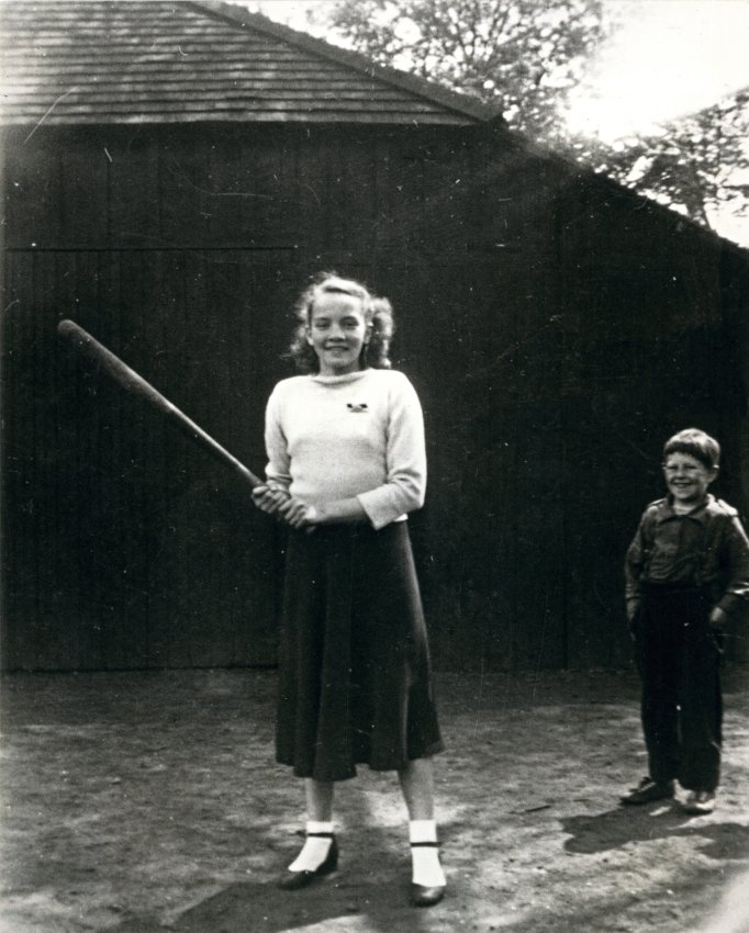 Black and white photographic reprint of Doreen Argue (married name is Doreen Moulton) outside the Victoria School on the playground. She is holding a baseball bat. She is wearing a white sweater with a pin, a long dark skirt, and socks and shoes. She has pigtails in her hair. A little boy is standing to the right with his hands in his pockets. Behind them is a wooden shed.