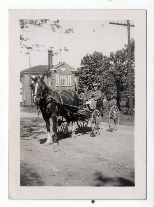 Rectangular black and white photograph of two women dressed in warm clothes seated in a horse-drawn carriage with the Victoria School (267 Edmund Street) in the background. There is a telephone pole on the right, and the road appears to be dirt.